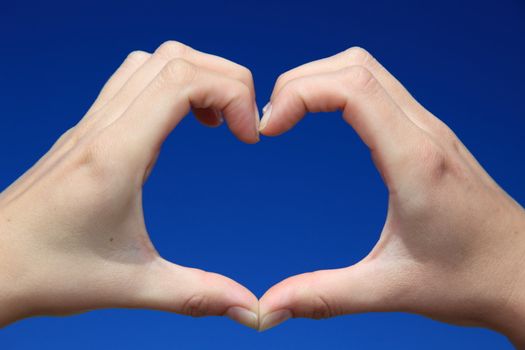 Persons hands forming a heart infront of bright blue sky.