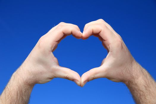 Persons hands forming a heart infront of bright blue sky.