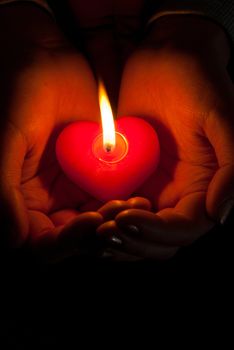 Human hands hold heart shaped burning candle against dark background