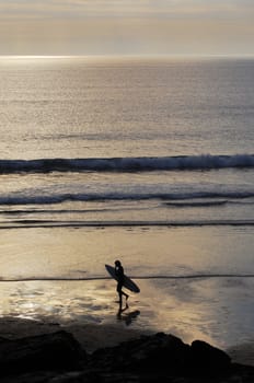 Surfer walking accross the beach at sunset. Trebarwith Strand, Cornwall, UK.