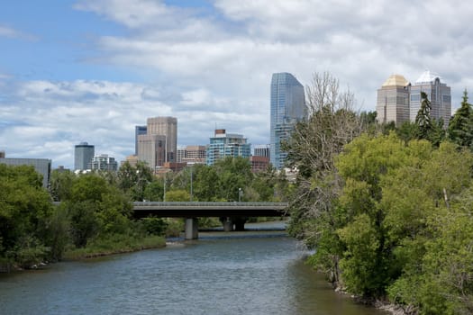 Blue skies reflected in the river with green trees in the foreground to the right and the city highrises towering over the scene.