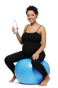 Full lenght portrait of a young smiling pregnant woman sitting on a blue ball, holding a bottle of water, isolated on a white background.