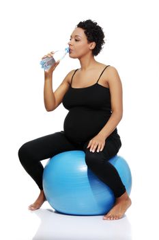 Full lenght portrait of a young smiling pregnant woman sitting on a blue ball, taking a sip of water, isolated on a white background.