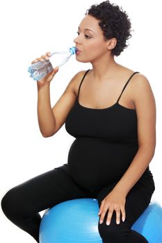 Closeup of a young smiling pregnant woman sitting on a blue ball, taking a sip of water, isolated on a white background.