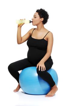 Full lenght portrait of a young smiling pregnant woman sitting on a blue ball, drinking an energy potion, isolated on a white background.