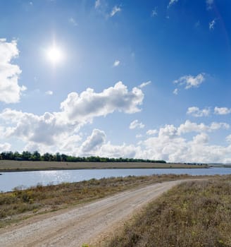 sun on cloudy sky over rural road and river near it