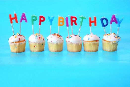 Cupcakes with Happy Birthday Candles on them against a blue background and plenty of room for copy space.