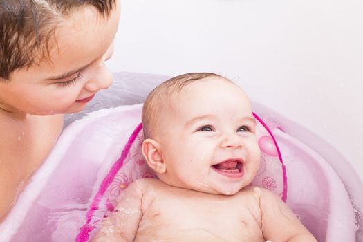Pretty smiley baby girl is having a bath in her bathtub with her, brother.
