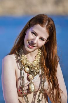 A beautiful woman with pale white skin and long red hair sitting on a rock semi-nude at the beach