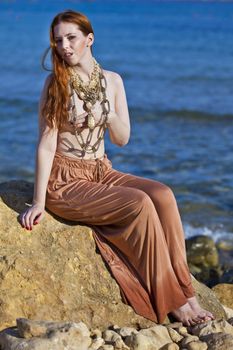 A beautiful woman with pale white skin and long red hair sitting on a rock semi-nude at the beach