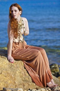 A beautiful woman with pale white skin and long red hair sitting on a rock semi-nude at the beach