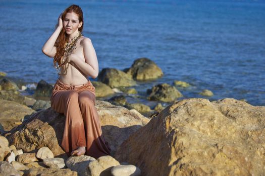 A beautiful woman with pale white skin and long red hair sitting on a rock semi-nude at the beach