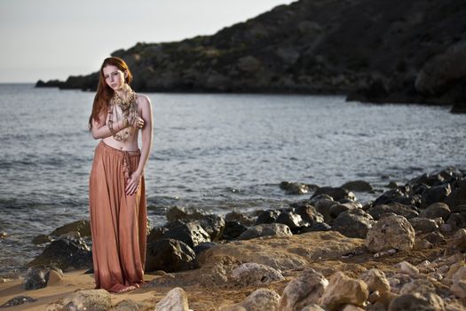 Beautiful woman with pale skin and long red hair wearing Bohemian jewellery and posing semi-nude on a beach