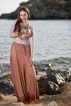 Beautiful woman with pale skin and long red hair wearing Bohemian jewellery and posing semi-nude on a beach