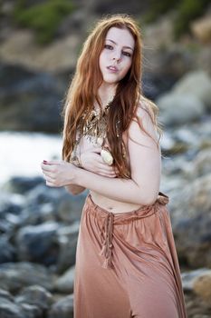 Beautiful woman with pale skin and long red hair wearing Bohemian jewellery and posing semi-nude on a beach