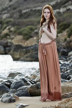 Beautiful woman with pale skin and long red hair wearing Bohemian jewellery and posing semi-nude on a beach