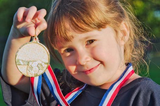 Little girl holding a gold medal with a soccer pattern at sunset