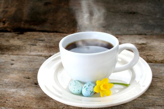 Cup of coffee with steam showing, also has Easter eggs and a daffodil on the saucer and shot on an old piece of wood for a rustic look.