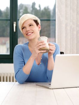 Photo of a beautiful blond female working in an office on a laptop and drinking a coffee.