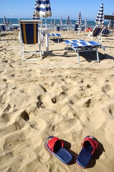 A pair of sandals on the beach with empty deck chairs and parasols in the background 