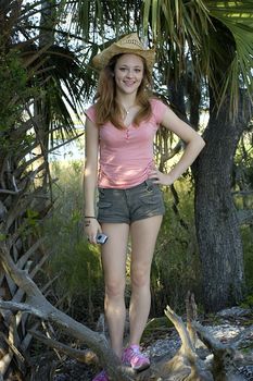 A girl poses on a piece of driftwood in her straw hat on the beach.