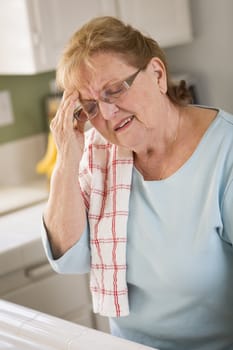 Grimacing Senior Adult Woman At Kitchen Sink With Head Ache.