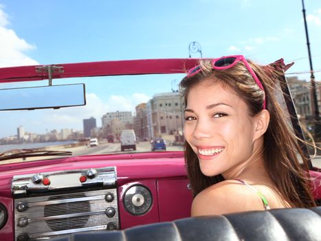 Car woman smiling happy on passenger seat in pink vintage convertible car. Young mixed race Asian / Caucasian female model during cuba vacation. Driving on Malecon waterfront, Havana, Cuba