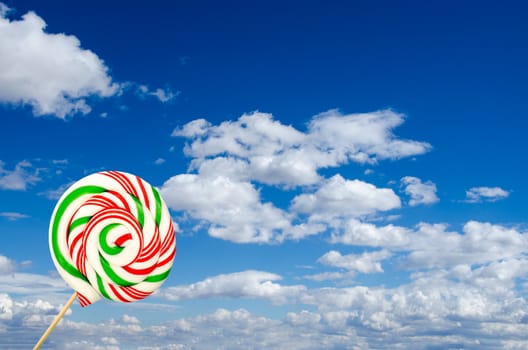 Single sugar lollipop in white green and red on background of sky and clouds