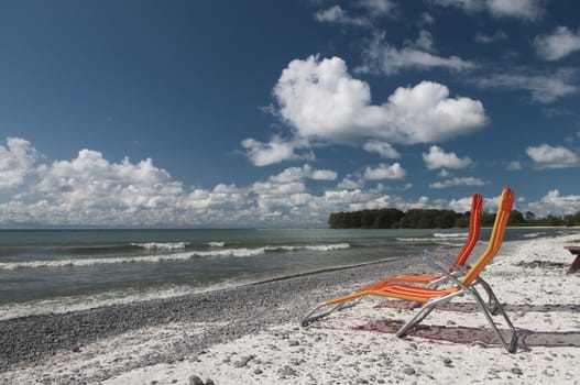 Two lounging chairs on the beach on Lake Ontario with fluffly white clouds on a deep blue sky