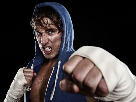 Boxer - street fighter in hoody fighting aggressive looking angry at camera hitting with hand wraps tape on hands. Young caucasian male fitness model isolated on black background.