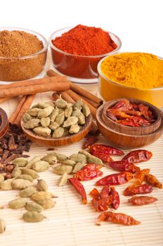 gorgeous setting with cooking spices and herbs (cloves, cardamom pods, cinnamon sticks, garam masala, paprika, piri piri, turmeric) on a wooden mat (isolated on white background)