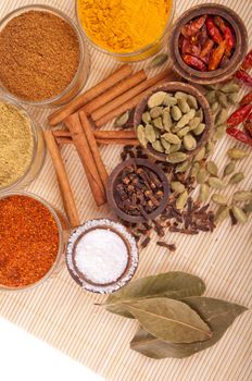 gorgeous setting with cooking spices and herbs (bay leaves, chili powder, coriander, cloves, cardamom pods, cinnamon sticks, garam masala, piri piri, salt, turmeric) on a wooden mat