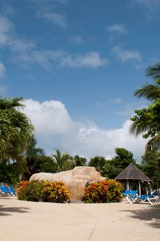 gorgeous swimming pool area with big artificial stone, chairs and bungalow on a tropical resort (surrounded by palm trees)