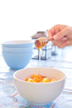 A woman's hand pouring clear honey onto a bowl of fresh fruit salad