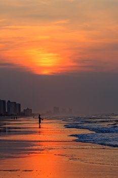 A silhouette of a lone fisherman fishing in the early morning in the Atlantic Ocean from the beach as the sun rises.