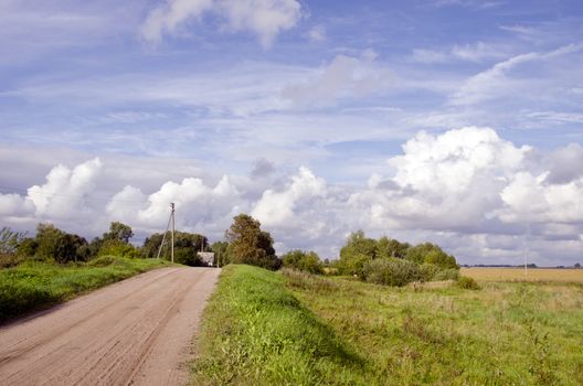 Rural gravel road and house in distance. Cloudy sky and landscape of meadows. Power poles.