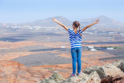 Young girl standing on cliff's edge and looking to a sky with raised hands