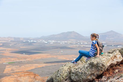 Young girl sitting with backpack on cliff's edge and looking to a sky. Lanzarote