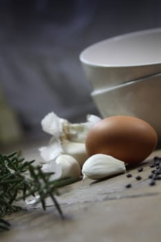 Close up detail of some kitchen ingredients comprising of a brown egg, garlic, fresh rosemary and black lentils. All set on a portrait format against a wooden background. Copy-space available.