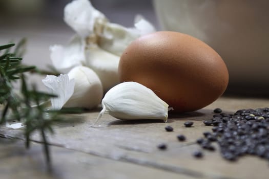 Close up detail of some kitchen ingredients comprising of a brown egg, garlic, fresh rosemary and black lentils. All set on a landscape format against a wooden background.