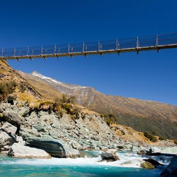 Simple suspension swing bridge crossing high over a rocky glacial mountain river in Mount Aspiring National Park South Island New Zealand