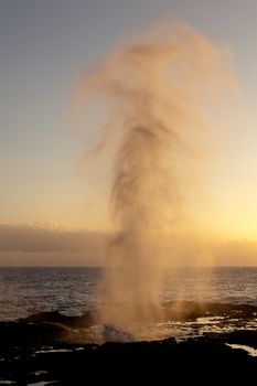 Setting sun illuminates spout of water from lava rocks on coast of Kauai