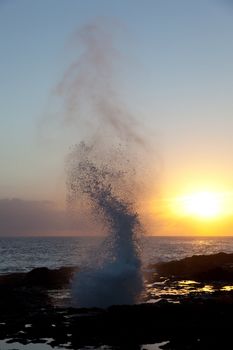 Setting sun illuminates spout of water from lava rocks on coast of Kauai