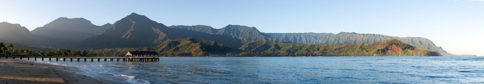 Rising sun illuminates the Hanalei pier and Na Pali mountains in Kauai in panoramic