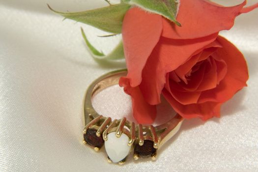 A macro of an opal and ruby ring with a tiny rosebud lying on white silk.