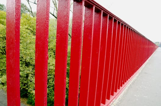 Bridge railing painted red background closeup. Man goes in distance and trees.