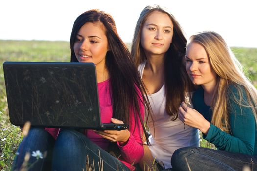 three young beautiful woman with a laptop sitting in the field on sky background 