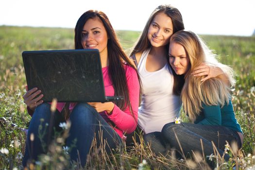 three young beautiful woman with a laptop sitting in the field on sky background 