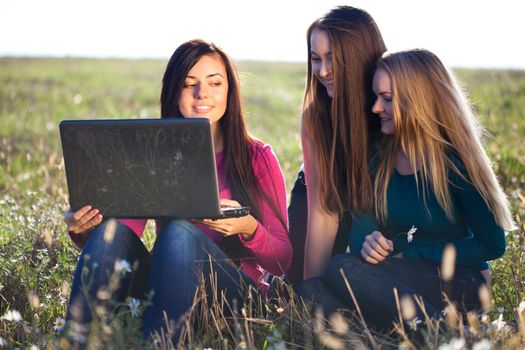 three young beautiful woman with a laptop sitting in the field on sky background 