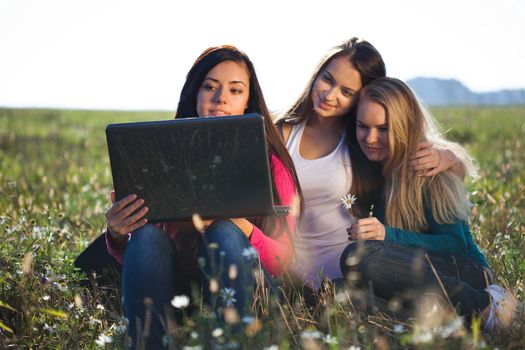 three young beautiful woman with a laptop sitting in the field on sky background 